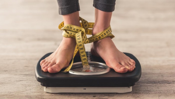 Cropped image of woman feet standing on weigh scales, on gray background. Legs winded with a tape measure