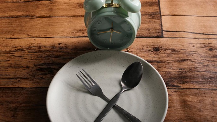 Alarm clock on a plate, surrounded by a burlap napkin and silverware, placed on a rustic wooden table. Intermittent fasting, ketogenic dieting, weight loss, meal planning, and healthy eating concepts