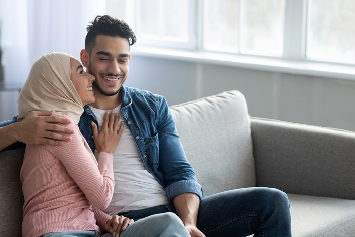 Happy middle-eastern family husband and wife cuddling on couch at home, embracing, talking and laughing, panorama with copy space. Cheerful arab couple enjoying time together