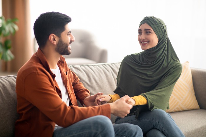 Happy Middle Eastern Couple Holding Hands Smiling To Each Other Sitting On Couch Spending Time Together At Home. Happiness In Relationship And Marriage Concept. Selective Focus