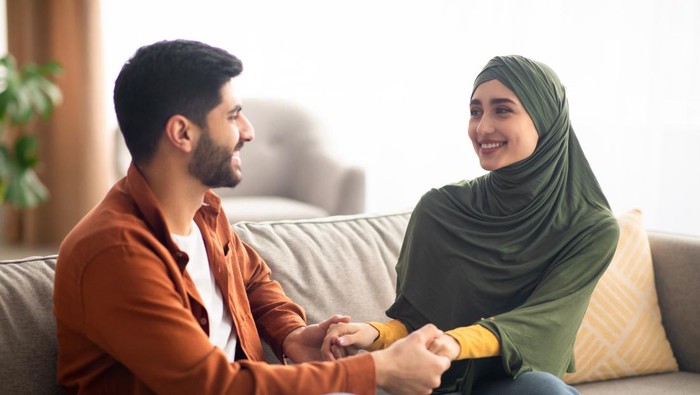 Happy Middle Eastern Couple Holding Hands Smiling To Each Other Sitting On Couch Spending Time Together At Home. Happiness In Relationship And Marriage Concept. Selective Focus