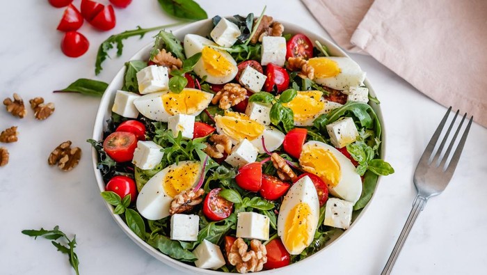 Angled view of a bowl of salad with hard-boiled egg, feta cheese, walnuts and cherry tomatoes, drizzled with olive oil