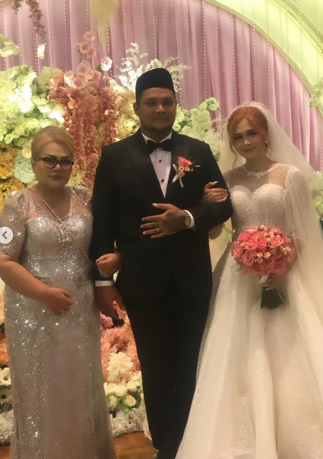 Ibunda Virgoun, Eva Manurung, memilih kenakan dress panjang abu-abu dengan model bagian atas sheer. Ia berpose bertiga bersama kedua mempelai. Foto: Instagram/@virgoun_/@lindifitriyana