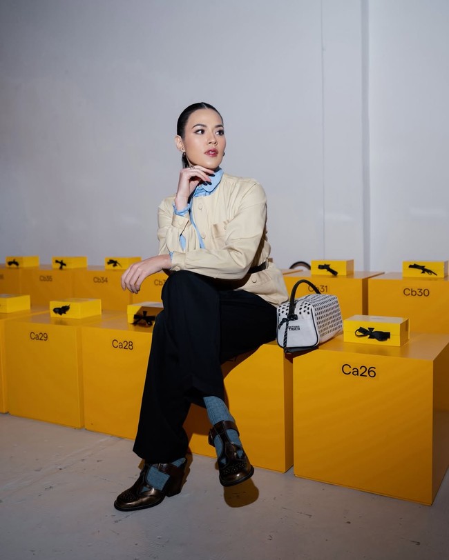 Ibu satu anak ini membawa tas tangan putih dengan detail stud metalik yang memberi aksen edgy. Sabuk hitam tipis di pinggang mempertegas siluet, sementara sepatu loafers cokelat dengan kaus kaki abu-abu menghadirkan nuansa smart-casual. Foto: Instagram/@raisa6690