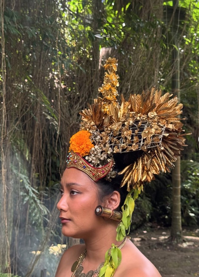 Salah satu yang tak kalah mencuri perhatian adalah mahkota besar berwarna emas yang dikenakan Tara. Hiasan kepala tersebut dipenuhi ornamen bunga dan detail ukiran khas Bali. Dipadukan dengan anting besar serta kalung emas bertumpuk, tampilannya semakin dramatis. Foto: Instagram/@tarabasro