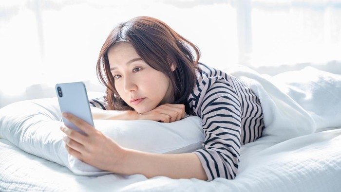 A woman falling asleep staring at a smartphone screen in bed.