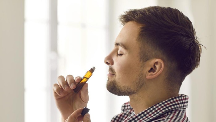Profile portrait of happy man enjoying aromatherapy. Side view of handsome young Caucasian man holding roller bottle and smelling fresh masculine scent of natural essential oil