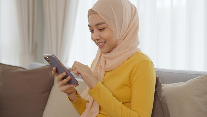 Young beautiful Muslim woman wearing hijab headscarf is using smartphone and selfie sitting on sofa at home.