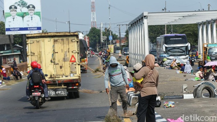 Penyapu koin membawa anak sambil berharap mendapatkan uang dari pengendara di Jembatan Kali Sewo, Pantura, Sabtu (14/3/2026). Aktivitas duduk di tepi jalan sambil membawa sapu itu sudah nampak sejak pagi hari. Mereka mengharapkan lemparan uang baik koin maupun kertas dari para pengendara yang melintas.