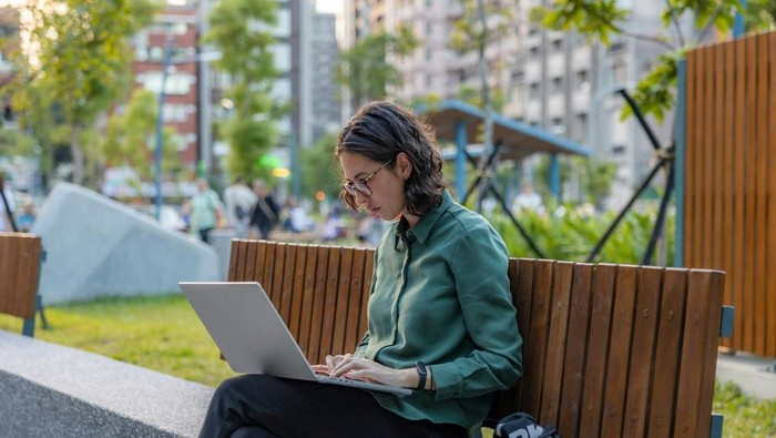 Seorang wanita duduk di taman sembari memangku laptop. Ilustrasi WFA.