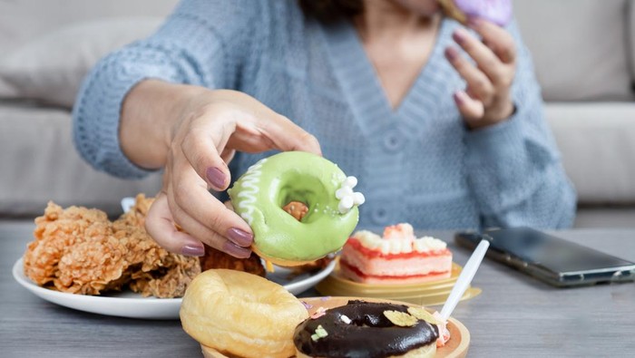 Woman reaching for a donut from a table full of junk food, showing signs of sugar cravings, overeating, and unhealthy eating habits