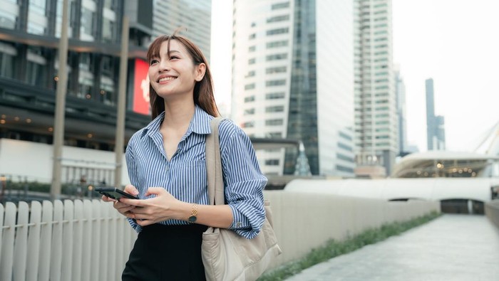 Happy young businesswoman walking to work, texting on her smartphone as she navigates the morning rush, confirming meeting agendas, reading real-time updates, and syncing with her remote team, balancing efficiency with ease, embodying the rhythm of m