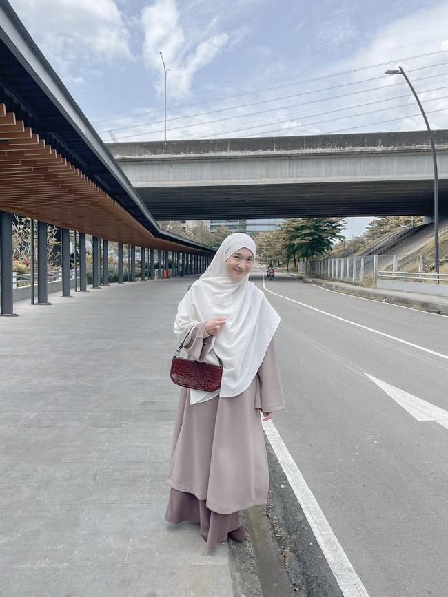 Tampil manis dengan setelan split abaya warna cokelat dan hijab syari warna putih, memberikan kesan yang tenang dan rapi. Penggunaan shoulder bag kecil dengan motif tekstur memberikan sentuhan modern pada padu padan busana yang santun. Foto: Dok. Instagram @denisarani.