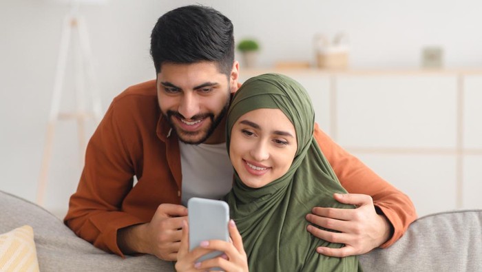 Happy Middle Eastern Spouses Using Mobile Phone Texting And Websurfing Sitting On Couch At Home. Modern Communication And Technology, Great Application For Cellphone Concept