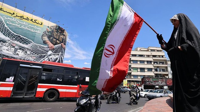 Seorang perempuan mengibarkan bendera nasional Iran di depan papan reklame raksasa bertuliskan Selat Hormuz tetap ditutup di Lapangan Revolusi, Teheran. Minggu (12/04). (Anadolu via Getty Images)