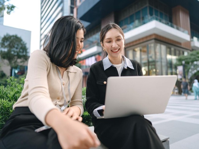 Positive laughing two asian businesswoman working friendship concept. Business women people enjoy talking using laptop. Modern technology lifestyle. Authentic candid sitting at outdoors with city building background.