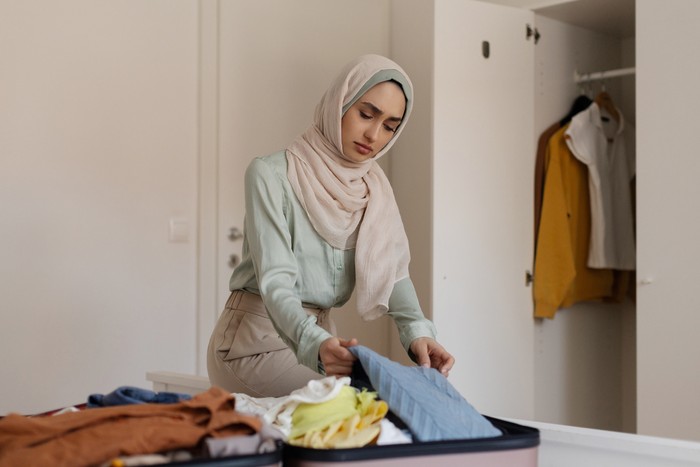 Young depressed Muslim woman packing clothes in suitcase in a small bedroom, lady looks worried and deep in thought