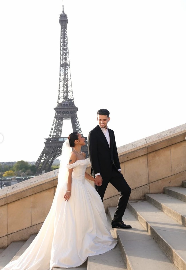 Rok gaun dibuat mengembang dengan ekor panjang yang menjuntai, menciptakan efek megah saat difoto di tangga dengan latar belakang Menara Eiffel. Ia melengkapi tampilannya dengan veil transparan panjang yang jatuh halus dari kepala, memberikan sentuhan klasik. Foto: Instagram/@jennifercoppenreal