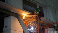 Lengan eskavator terlihat menghujam dengan presisi, lalu mematahkan bagian pilar serta balok beton di lantai dua fasad. Strategi ini dilakukan untuk melemahkan struktur atas terlebih dahulu sebelum nantinya didorong jatuh ke arah jalan raya yang sudah dipasangi barikade water barrier.