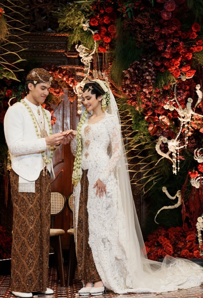 El tampil dengan beskap putih yang clean dan klasik, dipadukan dengan kain batik cokelat bermotif tradisional sebagai bawahan. Melengkapi penampilannya, El juga mengenakan aksesori khas pengantin pria Jawa, seperti blangkon bermotif senada dengan kain batiknya serta untaian melati (kalung melati) yang dikenakan di leher. Foto: Instagram/@syifahadju