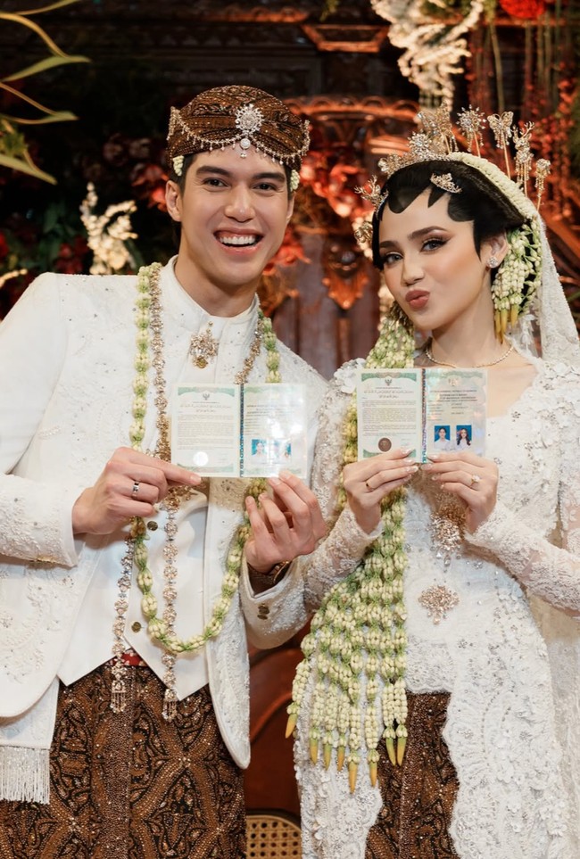 Syifa mengenakan kebaya pengantin berwarna putih dengan detail bordir dan payet yang halus. Siluetnya fitted mengikuti tubuh, dengan lengan panjang transparan yang dihiasi sulaman elegan. Bagian bawahnya memanjang membentuk train yang dramatis namun tetap lembut, menambah kesan mewah tanpa berlebihan. Foto: Instagram/@syifahadju