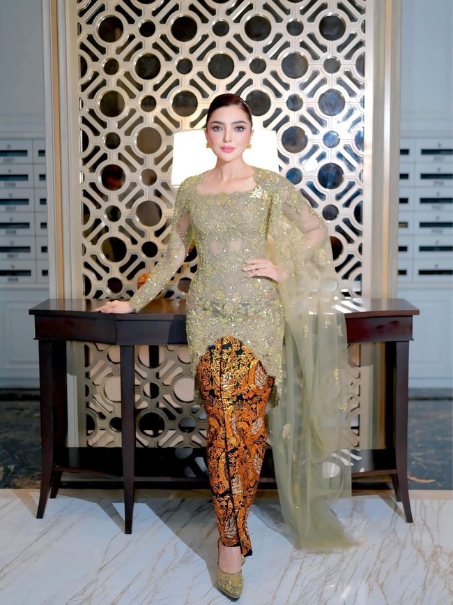 Foto ini menampilkan pose full-body Ashanty yang menonjolkan keindahan kebaya modifikasi dengan selendang tule transparan yang menjuntai. Kombinasi warna olive dan emas memberikan kesan mewah dan sophisticated. Dan aku suka karena ada hijaunya, ada tembaganya, terus warnanya itu kayak menampilkan warna baru yang aku belum pernah pakai juga gitu. Jadi aku suka warnanya, ucapnya. Foto: Dok. Instagram @ashanty_ash.