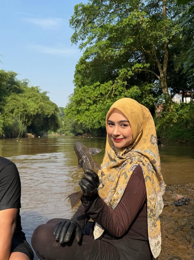 Saat terjun ke sungai untuk menangkap ikan sapu-sapu, Ricis tampil mengenakan baju cokelat dan jilbab motif warna kuning. Dia juga memakai sepatu boot untuk melindungi kakinya. Foto: Dok. Instagram Ria Ricis