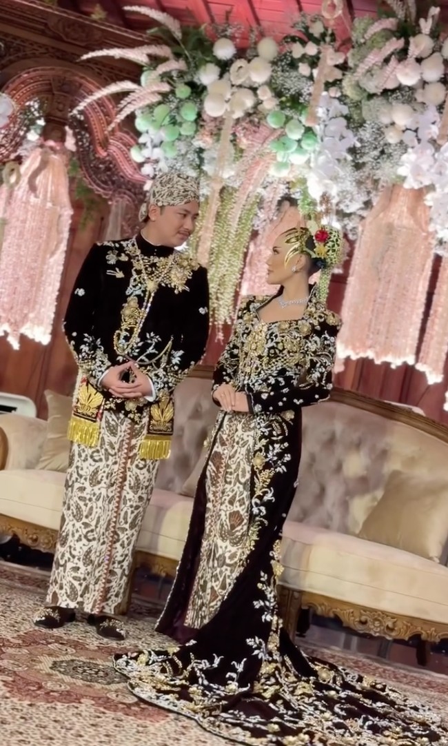 Untuk mempelai wanita, kebayanya dibuat dengan siluet fit body yang mempertegas bentuk tubuh, lalu dilengkapi ekor panjang dramatis (long train) yang menjuntai lebar di lantai. Material velvet warna coklat gelap memberi kesan elegan dan royal. Hampir seluruh permukaan kebaya dipenuhi bordir floral warna emas dengan detail beadwork dan kristal yang dijahit tangan, menciptakan efek berkilau saat terkena cahaya. Foto: Instagram/@ivan_gunawan
