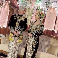 Untuk mempelai wanita, kebayanya dibuat dengan siluet fit body yang mempertegas bentuk tubuh, lalu dilengkapi ekor panjang dramatis (long train) yang menjuntai lebar di lantai. Material velvet warna coklat gelap memberi kesan elegan dan royal. Hampir seluruh permukaan kebaya dipenuhi bordir floral warna emas dengan detail beadwork dan kristal yang dijahit tangan, menciptakan efek berkilau saat terkena cahaya. Foto: Instagram/@ivan_gunawan