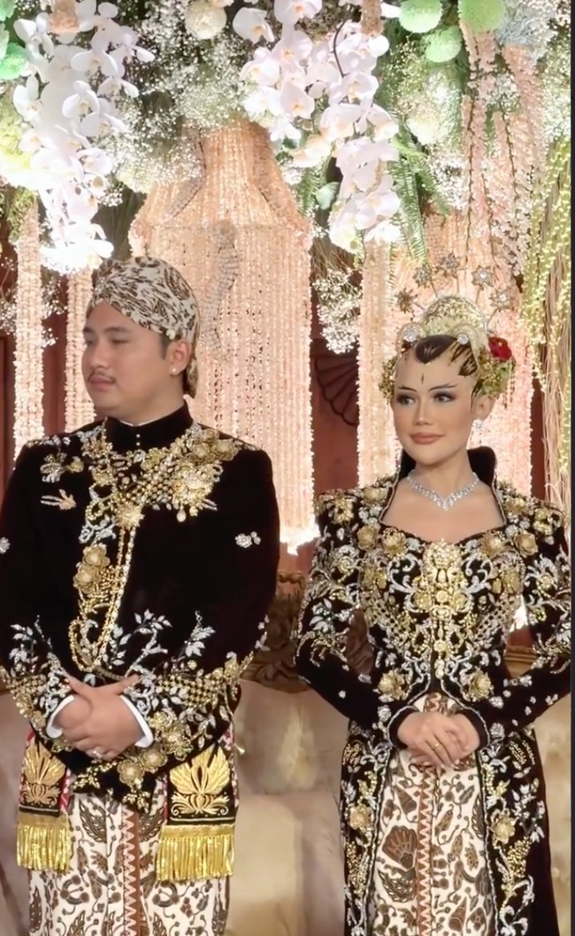 Sementara itu, mempelai pria mengenakan beskap velvet senada dengan detail bordir emas dan perak di bagian dada serta lengan. Ornamen bordir bernuansa floral dan klasik Jawa membuat tampilannya terlihat berwibawa. Beskap dipadukan dengan kain batik sogan tradisional bermotif besar dalam warna cokelat-krem khas Jawa. Foto: Instagram/@ivan_gunawan