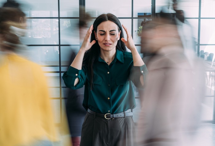Shot of overwhelmed businesswoman holding her head while working in a chaotic office environment with blurred colleagues rushing around. Stressed businesswoman experiencing a headache while standing in a busy modern office environment with blurred colleagues moving around in the background.
