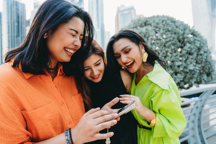 Three cheerful young women enjoying a sunny day in Dubai, laughing together while browsing a smartphone and sharing joyful moments