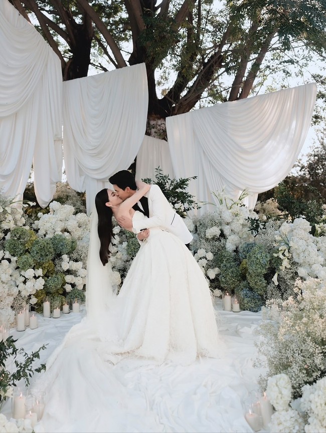 Tema all-white wedding kini tak lagi terlihat polos atau monoton. Dengan permainan tekstur bunga, layering dekorasi, hingga pencahayaan yang tepat, dekorasi putih bisa terlihat sangat mewah dan dreamy, seperti pernikahan pasangan aktris-aktor Thailand, Davika Hoorne dan Chantavit Dhanasevi. Foto: Instagram