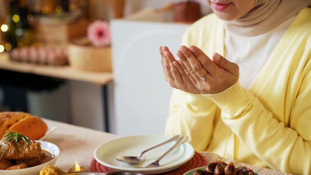 Muslim woman in hijab raises her hands in prayer before enjoying an iftar meal at home. Quiet ritual reflects devotion, gratitude, and the meaningful pause observed before breaking fast during Ramadan evening.