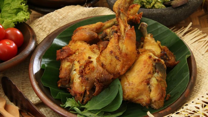 Ayam Penyet Sambal Hijau, a popular main dish in Surabaya, East Java. Marinated chicken pieces lightly smashed then deep fried; served with green chili paste, fried marinated tofu and tempeh, and fresh vegetables. The chicken, tofu, tempeh, and vegetables are served on traditional earthenware crockery while the green chili paste is served on a stoneware mortar. This is a close-up view of the fried chicken.