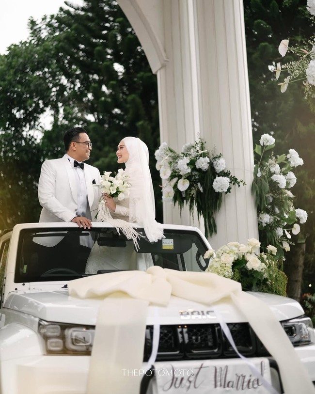 Dhani dan Nada tampil serasi saat berdiri di atas mobil Baic putih yang dihias pita besar, menciptakan momen wedding car yang modern, gagah, sekaligus tetap terlihat elegan. Foto: Dok. Instagram @thepotomoto.