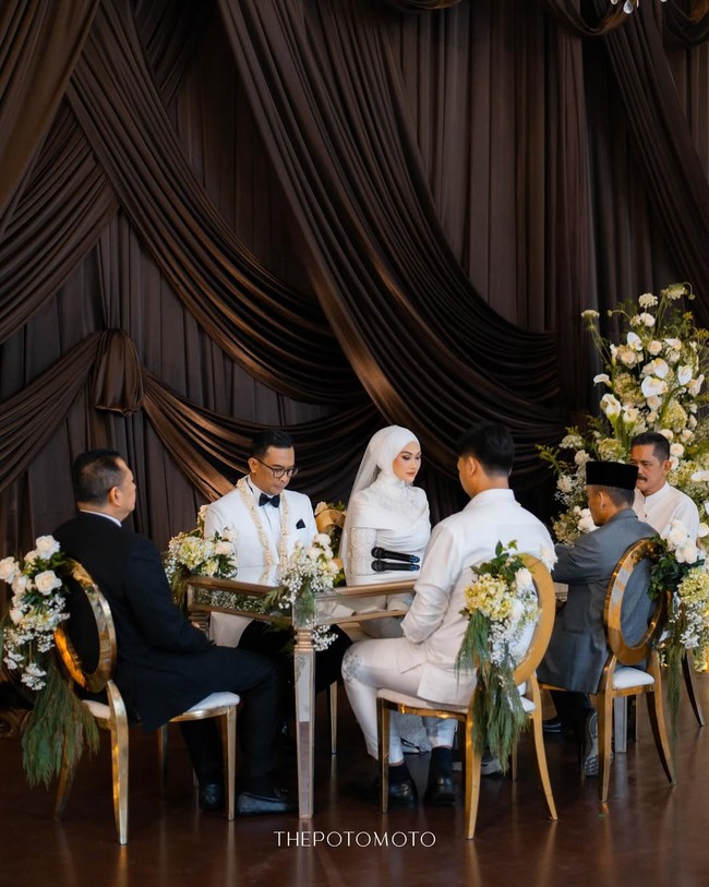 Pernikahan ini mengusung dekorasi bergaya klasik elegan dengan latar belakang tirai kain drapery berwarna cokelat tua yang megah, dipadukan dengan sentuhan romantis dari pencahayaan lilin hangat dan aransemen bunga putih yang anggun di sudut ruangan dari Artluxe Decor @artluxedecor. Foto: Dok. Instagram @thepotomoto.