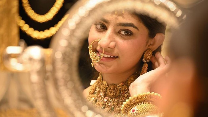 Ilustrasi. Seorang perempuan terlihat di cermin saat dia mengenakan kalung emas tebal, gelang, dan anting-anting, dengan berbagai perhiasan emas lain dipajang di latarnya. (Getty Images)