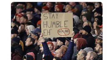 Suporter Arsenal membawa poster “Stay Humble” sebagai simbol pelampiasan setelah penantian gelar bertahun-tahun. Foto: X.com