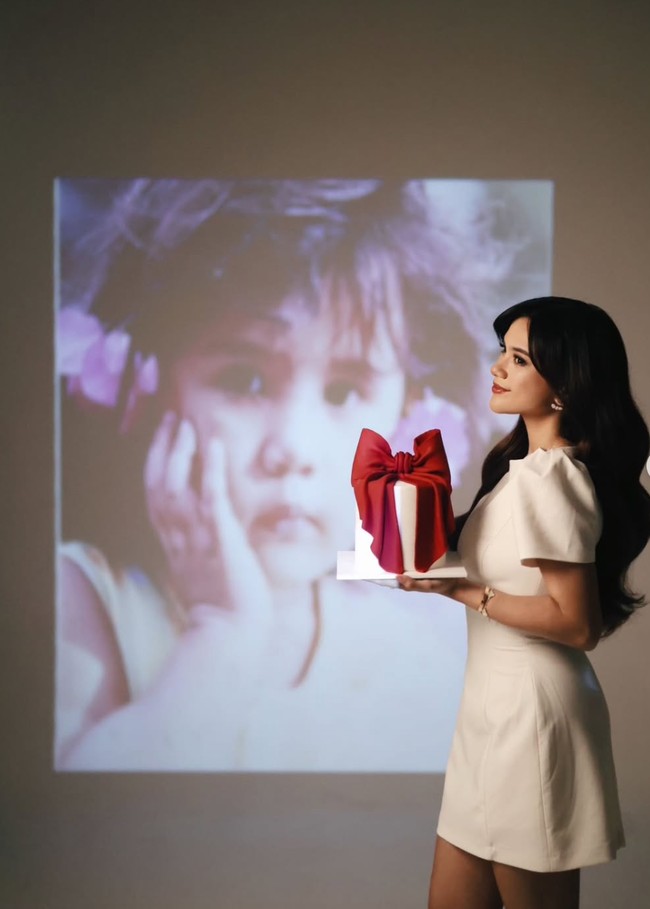 Dalam unggahan berbeda Audi terlihat berdiri di depan layar yang memperlihatkan fotonya saat kecil. Ia memegang kue ultah simpel berhias bentuk pita yang didominasi warna putih dan merah. Foto: Instagram/@audimarissa