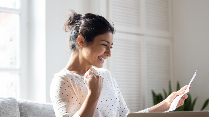 Happy Indian woman reading good news in letter, notification, holding paper, smiling girl rejoicing success, student excited by positive exam results, showing yes gesture, sitting on couch at home