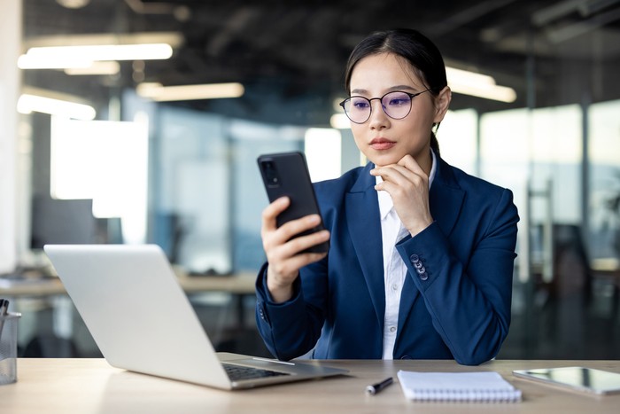 Asian businesswoman in office using smartphone while working on laptop. She wears glasses and blue suit, appearing focused and professional. Image conveys productivity, technology, modern work