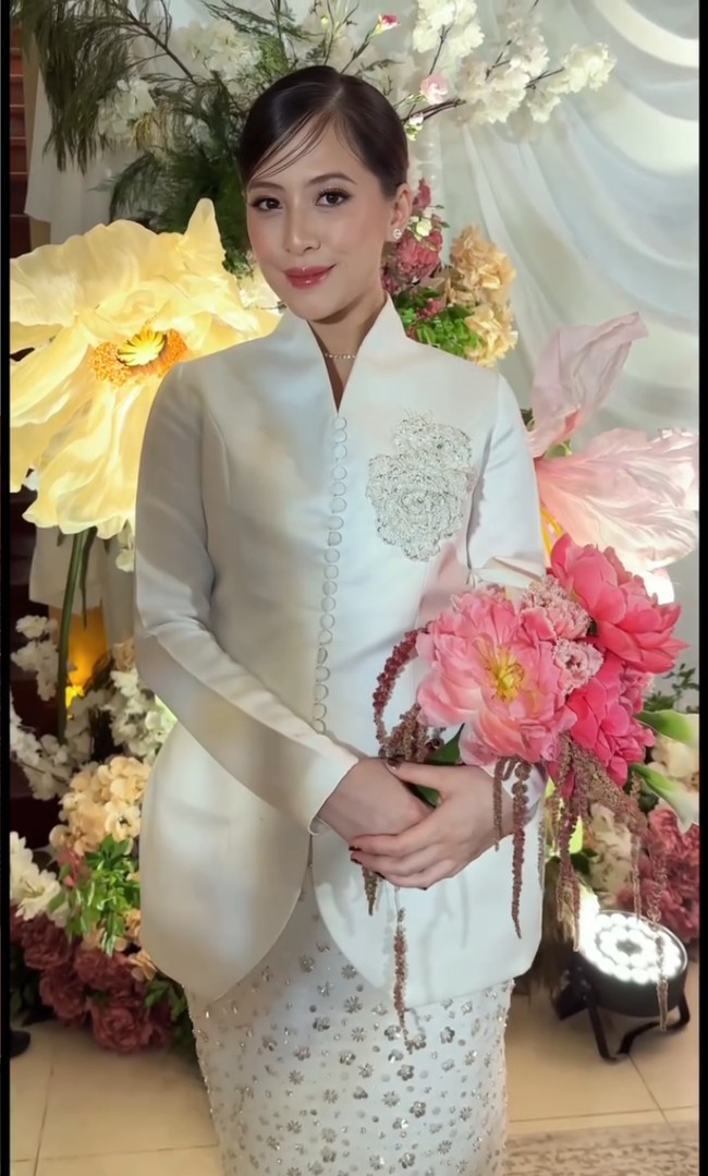 Kebaya putih datang dari koleksi Laviena Wedding Gallery. Kebaya kerah tinggi tampak manis berkat aksen mawar berpayet, berpadu apik dengan rok panjang berhias ratusan aplikasi bunga. Foto: Instagram/@anandachrstie