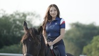 Dari atas punggung kuda hitamnya, Manda Cello melempar senyum manis ke arah kamera. Mengenakan kaos polo bernuansa biru dengan tambahan garis putih-merah yang dipadukan dengan celana berkuda gelap dan sepatu boots cokelat tinggi, penampilannya terlihat sangat sporty namun tetap elegan. (Foto: ist)