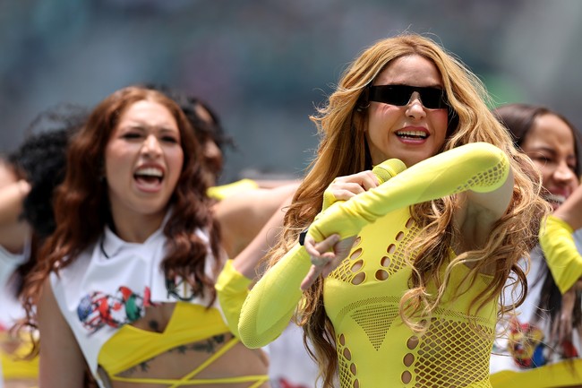 Lapisan pertama berupa fitted bodysuit dengan model mock-neck berwarna kuning neon yang mencolok. Siluet ini bukan hal baru bagi Shakira, yang kerap mengenakan one-piece saat tampil, termasuk dalam tur Las Mujeres Ya No Lloran tahun lalu. Potongan cut-out di bagian dada dan torso menambah dimensi pada busana yang ketat tersebut, dilengkapi sarung tangan opera tanpa jari dengan detail serupa. (Foto: Ryan Pierse/FIFA via Getty Images)