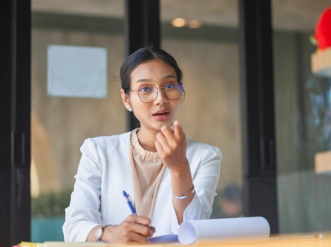 Confident young Asian businesswoman wearing glasses, presenting a project and gesturing during a professional meeting in a modern office, taking notes on documents with focused expression.