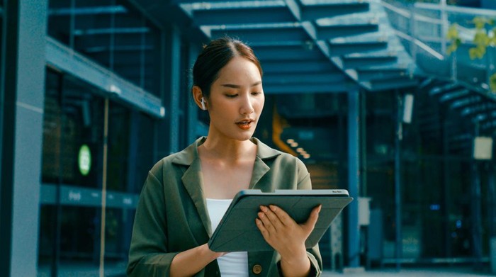 Closeup of young Asian businesswoman discussion walking and using digital tablet and wireless earphones outside modern office building. Remotely work and Sustainable business outdoor concept.