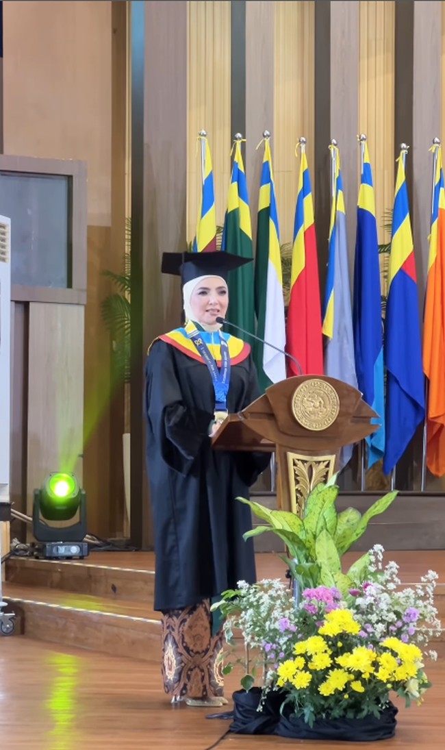 Dr. Asanti Hasuti (Ashanty) berdiri dengan anggun di podium untuk menyampaikan pidato kelulusannya sebagai wisudawan program Doktor. Berlatar deretan bendera universitas, momen ini menjadi pembuktian atas kerja keras dan ketahanan dirinya dalam menyelesaikan studi S3 di tengah kesibukan yang padat. Foto: Dok. Instagram @ashanty_ash.