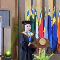 Dr. Asanti Hasuti (Ashanty) berdiri dengan anggun di podium untuk menyampaikan pidato kelulusannya sebagai wisudawan program Doktor. Berlatar deretan bendera universitas, momen ini menjadi pembuktian atas kerja keras dan ketahanan dirinya dalam menyelesaikan studi S3 di tengah kesibukan yang padat. Foto: Dok. Instagram @ashanty_ash.