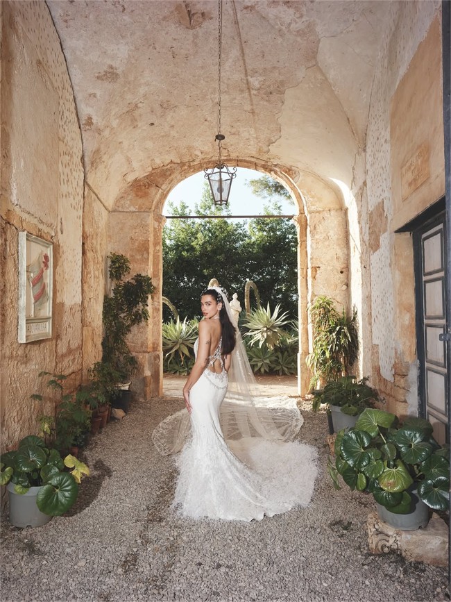 Siluetnya menampilkan potongan punggung rendah, lengkap dengan ekor gaun panjang yang dihiasi bulu-bulu. Penampilannya semakin dramatis berkat veil bordir panjang dan hiasan kepala berbulu. Foto: Instagram/@dualipa
